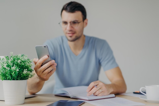 Moderne slimme telefoon in handen van de man Drukke mannelijke kantoormedewerker ontvangt berichten op de mobiele telefoon terwijl hij enkele aantekeningen maakt in het dagboek tegen een gezellig interieur Focus op cellulair technologieconcept