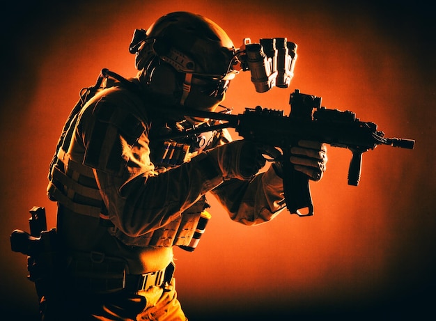 Moderne soldaat van speciale legertroepen, politie-antiterroristische squadronjager in gevechtsuniform, helm met nachtzichtapparaat gericht op een aanvalsgeweer met korte loop, rustige studio-opname met rode achtergrondverlichting