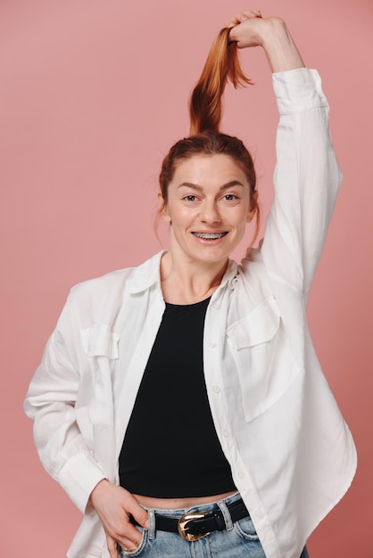 Moderne sportieve vrouw met rood haar met hemd en spijkerbroek die op roze achtergrond poseert