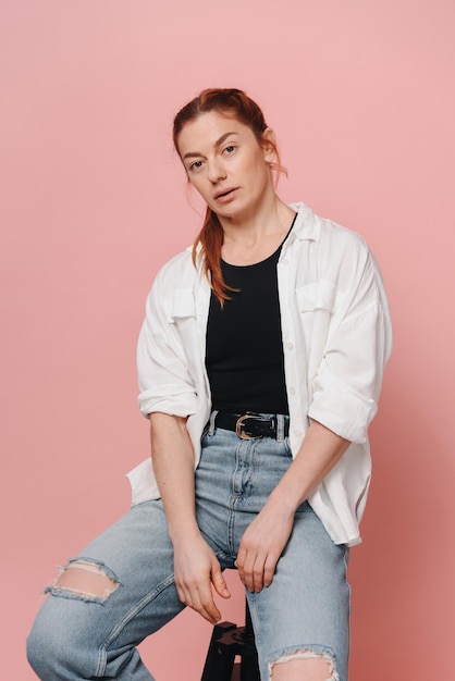 Moderne sportieve vrouw met rood haar met hemd en spijkerbroek die op roze achtergrond poseert