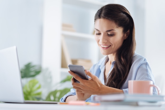 Moderne technologie. Blije positieve opgetogen vrouw aan de tafel zitten en glimlachen tijdens het gebruik van haar smartphone