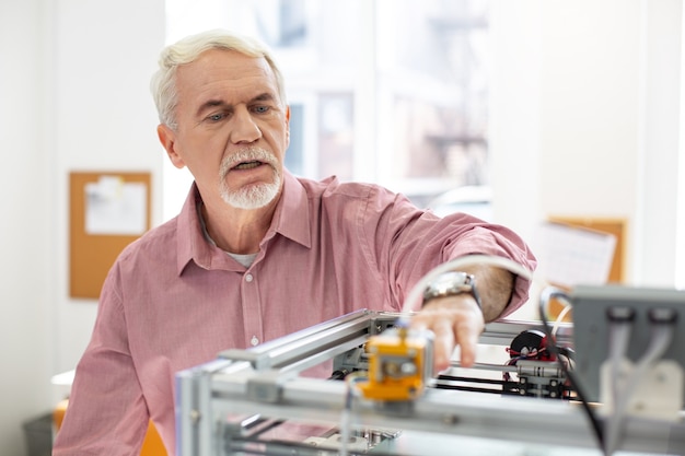 Moderne technologieën. Aangename senior man die een 3D-printer runt en iets afdrukt terwijl hij op kantoor werkt