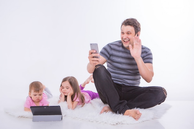 Moderne technologieën in het dagelijks leven: een man praat aan de telefoon via een headset, kinderen kijken naar een tekenfilm op een tablet. Hobby's en recreatie met gadgets. Ouder met meisjes op de vloer