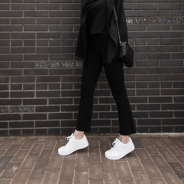 Moderne vrouw in modieuze denim broek in lederen stijlvolle sneakers in trendy jas met zwarte handtas staat in de buurt van bakstenen muur in de stad. Close-up vrouwelijk lichaam. Casual jeugdmodekleding voor dames.