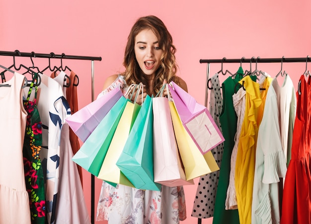 moderne vrouw permanent in de winkel in de buurt van kledingrek en houdt van kleurrijke boodschappentassen geïsoleerd op roze