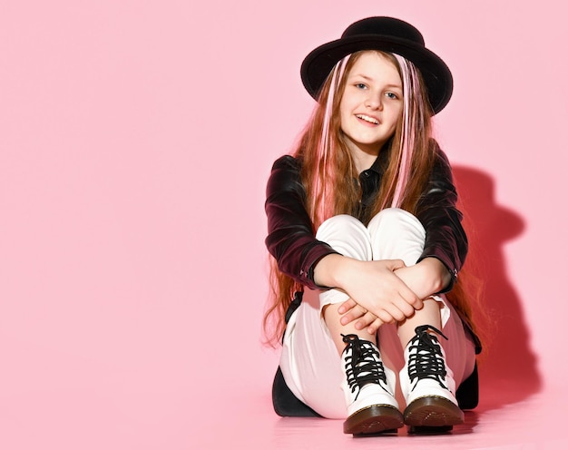 Modest teenage girl in a fashionable bow sitting on the floor with her legs tucked up posing on a pink background in a hat and black jacket