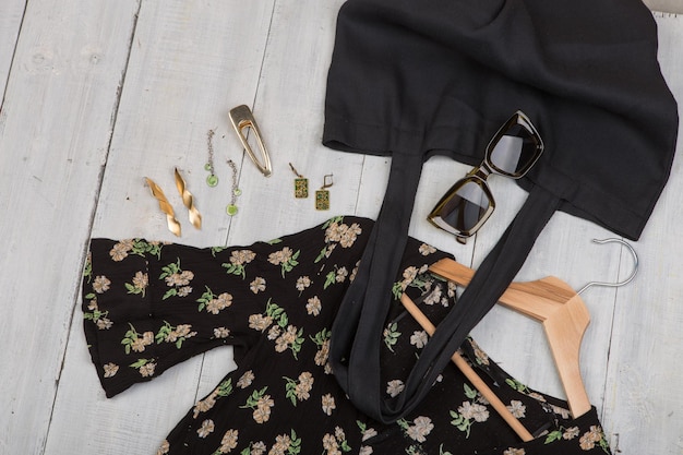 Modetrends - zonnebril, zwarte eco-draagtas en jurk in bloemenprint op hanger, sieraden: haarspeldje, zilveren en gouden oorbellen op wit houten bureau