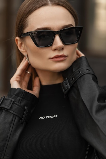 Modieus cool vrouwelijk portret van een mooi stijlvol hipstermeisje in een modieuze zwarte stedelijke outfit met modieuze zonnebril op straat