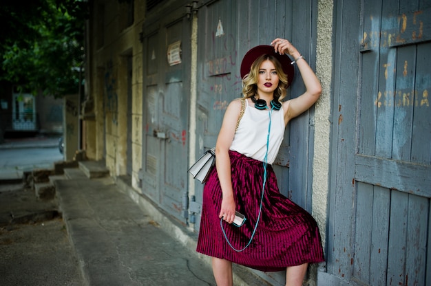 Modieus en mooi blond modelmeisje in een stijlvolle rode fluwelen velours rok, witte blouse en hoed, gesteld met telefoon en oortelefoons.