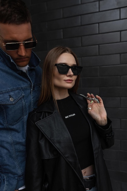Modieus jong schoonheidsmeisje met modebrillen in zwarte trendy outfit met tas staat in de buurt van een zwarte bakstenen muur met coole hipster-man