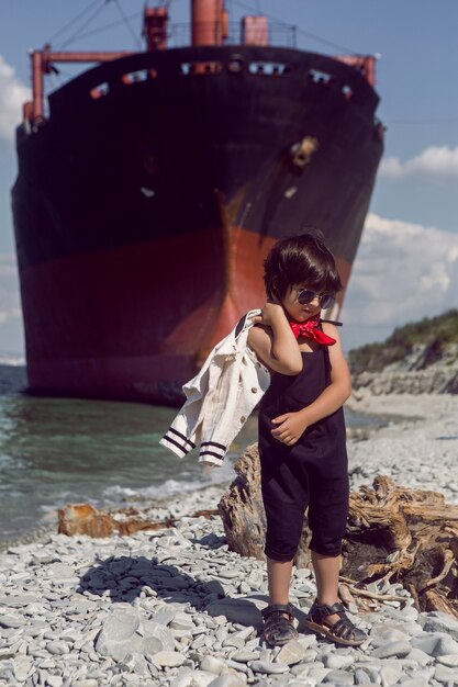 Modieus jongenskind met lang haar staat op een boomstam naast een groot schip dat bij Novorossiysk aan de grond liep