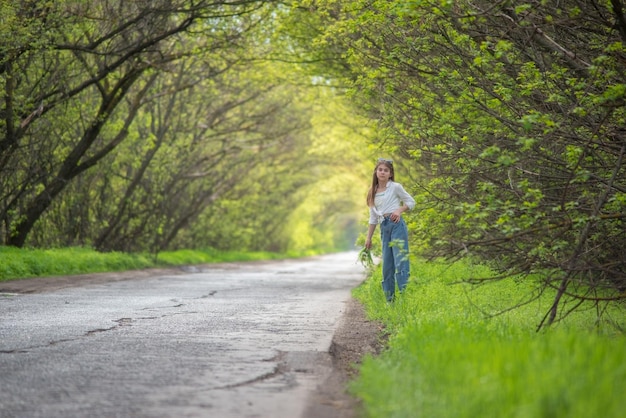 Modieus meisje dat op de weg loopt tegen de achtergrond van een boog van groene vertakte bomen