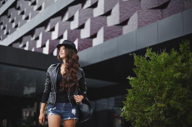 Modieus model meisje met golvend haar in een leren jas, zwarte hoed en jeans broek met een stijlvolle rugzak staat op straat in de buurt van modern gebouw. Trendy casual outfit. Elke dag kijken. Straatmode.
