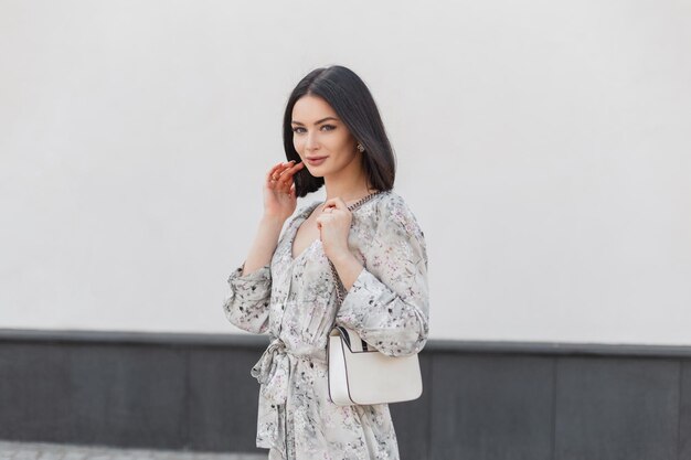 Modieus mooi brunette vrouw model met korte broek kapsel in stijlvolle vintage bloemen jurk met mode witte tas loopt in de buurt van een wit gebouw op straat