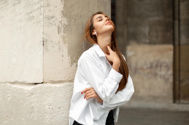 Modieus mooi elegant modelmeisje in stijlvolle kleding met een wit overhemd staat in de buurt van een betonnen vintage gebouw op straat