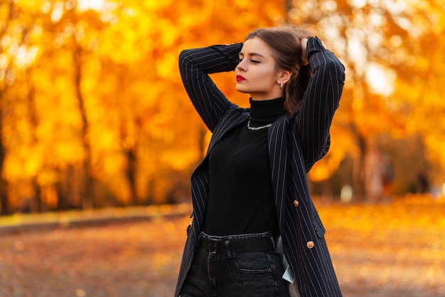 Modieus mooi meisje in een elegant pak met een stijlvol jasje met een zwarte trui en een leren rugzak op herfstnatuur met geel herfstgebladerte bij zonsondergang