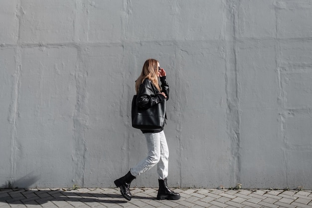 Modieus mooi meisjesmodel in jonge stijlvolle kleding met een leren jas, jeans en laarzen met een tas, zet een zonnebril op en loopt op een zonnige dag in de buurt van een grijze muur