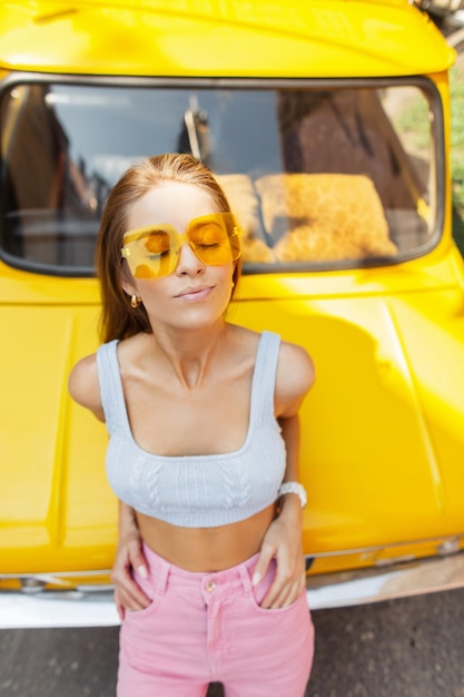 Modieus mooi stijlvol meisje met coole zonnebril en heldere zomerse trendy kleding geniet van een zonnige dag in de buurt van een vintage gele auto