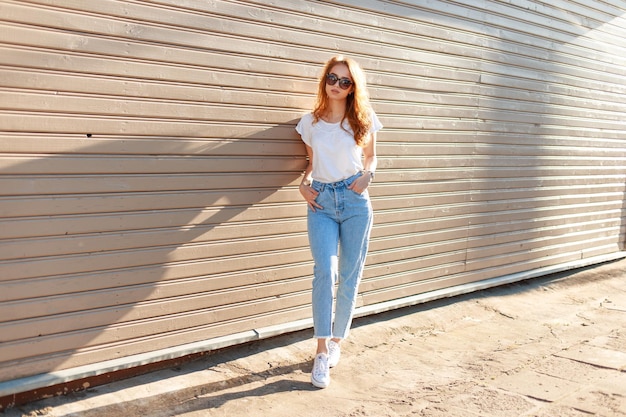 Foto modieus mooi stijlvol meisje model in casual kleding met een t-shirt en hoge taille jeans in de buurt van een vintage houten gebouw