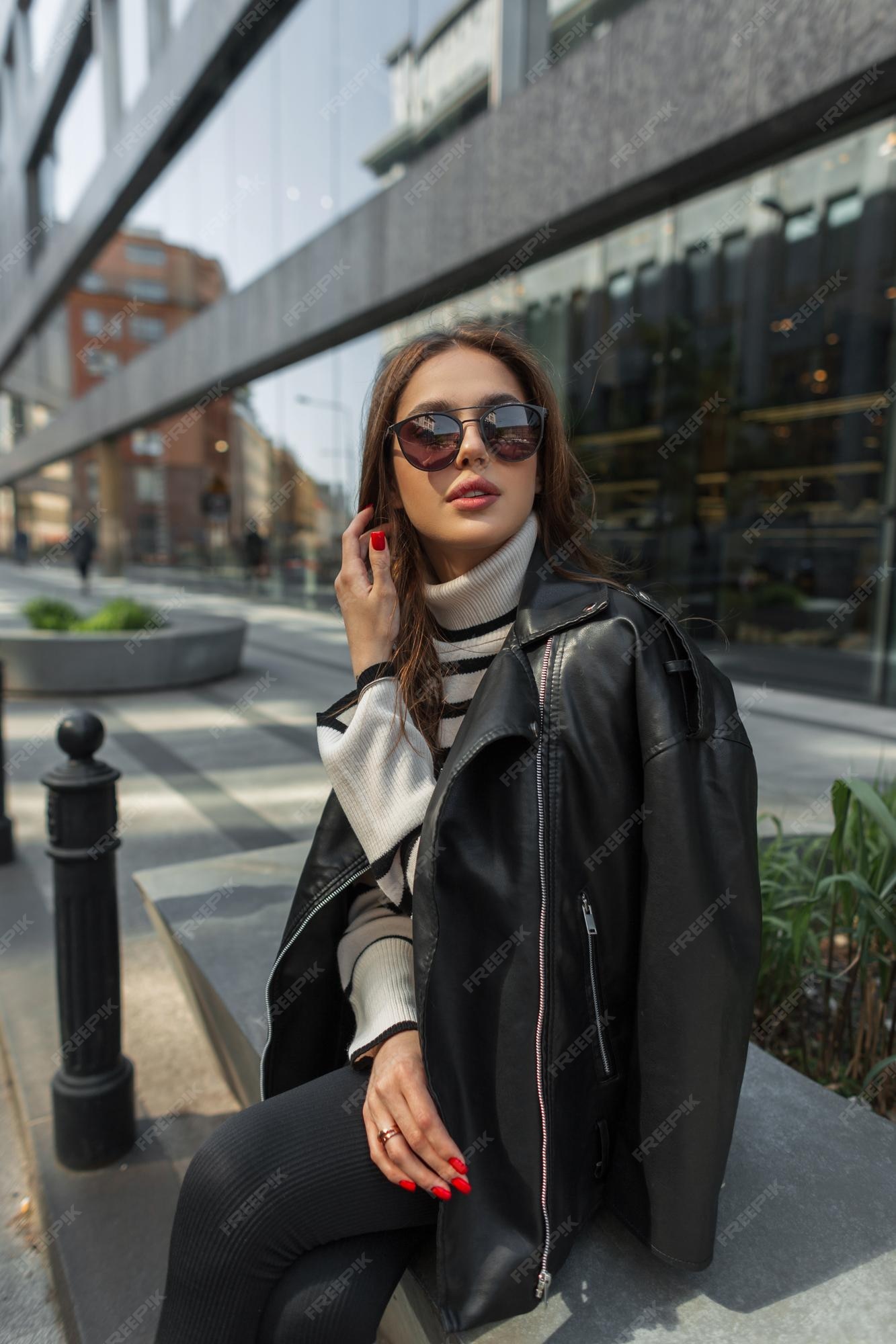 Oeps Minnaar Dij Modieus mooi trendy meisje met zonnebril in casual urban outfit met leren  jas en trui zit in de stad in de buurt van een modern gebouw | Premium Foto