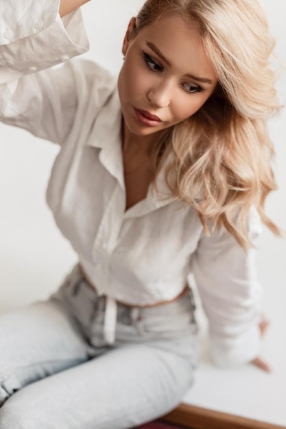 Modieus portret headshot van een mooi blond meisje met haar in een wit overhemd en spijkerbroek zittend in de studio