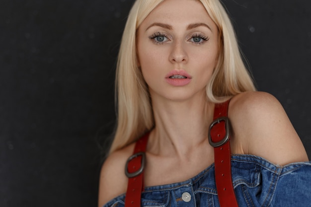 Modieus portret van een mooie jonge aantrekkelijke vrouw met sproeten en natuurlijke make-up in stijlvolle vintage denim kleding op een zwarte muur.