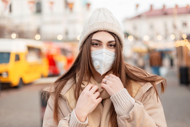 Modieus portret van een mooie jonge vrouw met een medisch beschermend masker in modieuze bovenkleding met een gebreide muts en jas die door de stad loopt