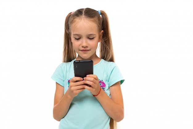 Modieus tienermeisje in blauw t-shirt communiceert met haar vrienden met telefoon en glimlacht op witte muur