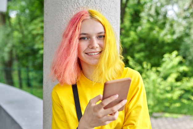 Modieus tienermeisje met gekleurd geverfd haar met een smartphone buitenshuis. Glimlachende heldere jonge vrouw in geel t-shirt met geel roze haar