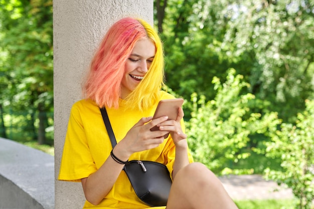 Modieus tienermeisje met gekleurd geverfd haar met een smartphone buitenshuis. Glimlachende heldere jonge vrouw in geel t-shirt met geel roze haar