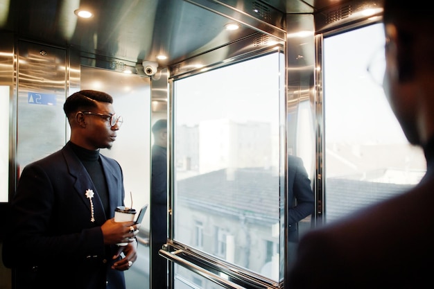 Modieuze Afro-Amerikaanse man in pak en bril met mobiele telefoon en kopje koffie bij handen geposeerd in lift