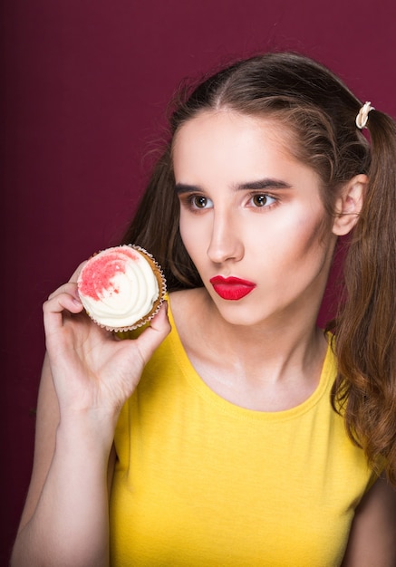 Modieuze donkerbruine vrouw die met lichte make-up zoete cupcake houdt. Studio-opname op een rode achtergrond