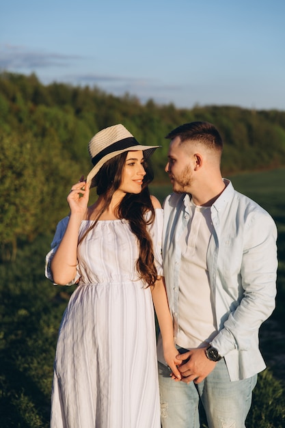 Modieuze en stijlvolle gelukkig zwangere vrouw en haar man gekleed een pastel wit en blauw toon in het veld op de zonsondergang