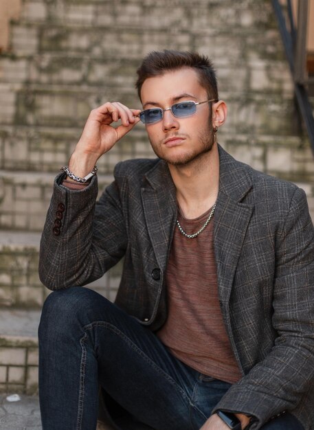 Modieuze hipster-man in een stijlvolle casual jas T-shirt en spijkerbroek zit op de vintage trappen en zet zijn stijlvolle blauwe zonnebril op