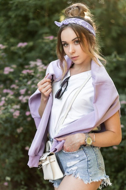 Modieuze hipster vrouw met een stijlvolle bandana in een wit t-shirt met een paarse hoodie en gescheurde denim shorts loopt in het park in de buurt van bloemen