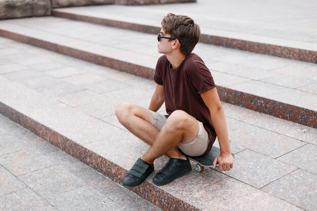 Modieuze jonge hipster man in zwarte zonnebril in een vintage t-shirt in trendy korte broek in sneakers rusten zittend op een skateboard in de stad op een zomerdag