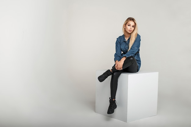 Modieuze jonge model vrouw in mode vintage jeanswear in lederen broek en stijlvolle sneakers zit op een wit vierkant in de studio