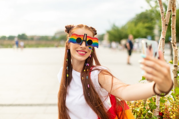 Modieuze lesbienne in kleurrijke zonnebril met lgbt regenboog op haar gezicht selfie te nemen met smartpthone op stadsplein