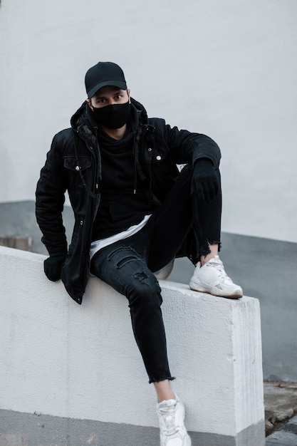 Modieuze man in zwarte stijlvolle kleding met zwarte design pet, jas, hoodie, jeans en witte sneakers met een beschermend masker buiten zitten