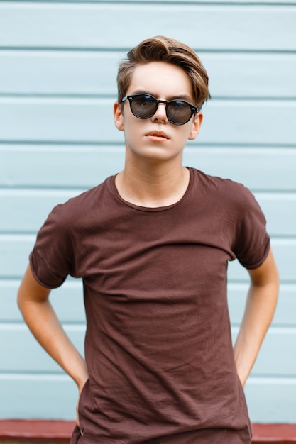 Modieuze moderne jonge hipster man in donkere zonnebril in trendy zomer t-shirt met stijlvol kapsel staat in de buurt van de vintage blauwe houten muur
