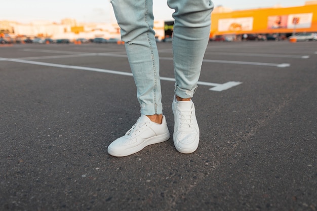 Modieuze moderne jongeman in stijlvolle blauwe spijkerbroek in een trendy lederen seizoensgebonden witte sneakers staat op een parkeerplaats in de stad. Close-up van de stijlvolle benen van mannen. Zomerse look.