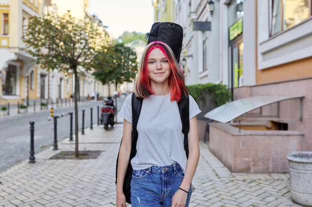 Modieuze, mooie, creatieve tienermeisjesmuzikant met gitaar voor het geval dat ze langs de stadsstraat loopt, glimlachende vrouw die naar de camera kijkt