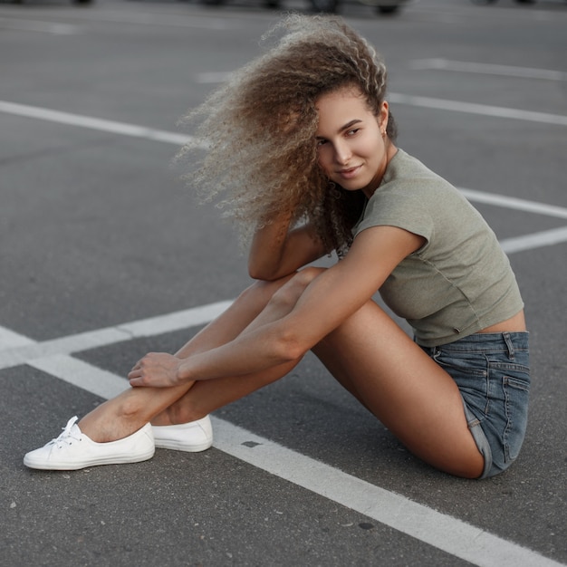 Modieuze mooie jonge gelukkig krullende vrouw in stijlvolle korte broek en witte schoenen zit op het asfalt