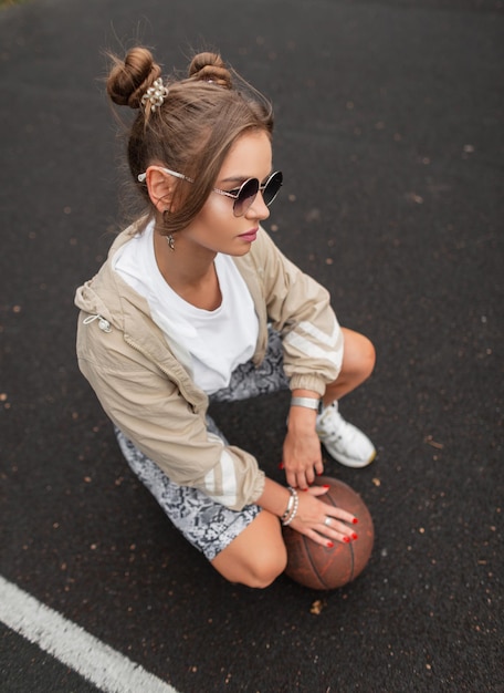 Modieuze mooie jonge hipster meisje met vintage zonnebril in stijlvolle kleding met een windbreaker tshirt legging en sneakers met een bal op het basketbalveld