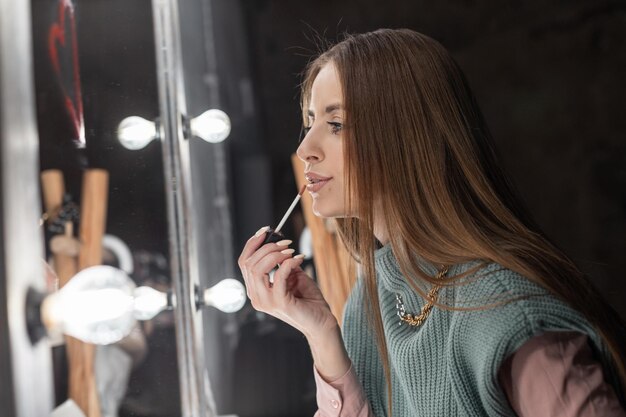 Modieuze mooie jonge vrouw in stijlvolle vintage kleding maakt make-up in de buurt van de spiegel en bereidt zich voor op een feestje Meisje schildert lippen met roze glans