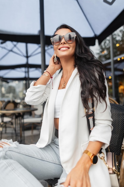 Modieuze mooie jonge zakelijke gelukkige vrouw met een glimlach met stijlvolle zonnebril in een modieuze witte blazer-topjeans met een handtas zit in een café in de stad