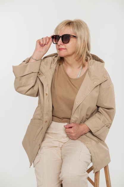 Modieuze mooie stijlvolle blonde oudere vrouw in trendy mode beige kleding met een windjack zet een zonnebril op en zit op een stoel op een witte achtergrond