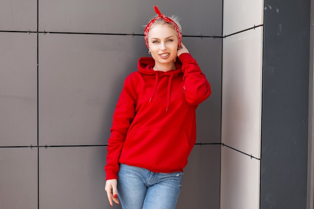 Modieuze vrolijke jonge vrouw met een schattige glimlach in moderne modieuze kleding met een rode bandana staat in de buurt van een grijs vintage gebouw op een warme zomerdag