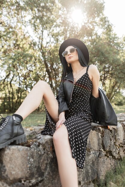 Modieuze vrouw brunette in fashion zonnebril in stijlvolle zwarte mooie kleding in zwarte vintage hoed ontspant op stenen op de natuur. Trendy meisje geniet van rust en lentezonlicht in het park in de lente.