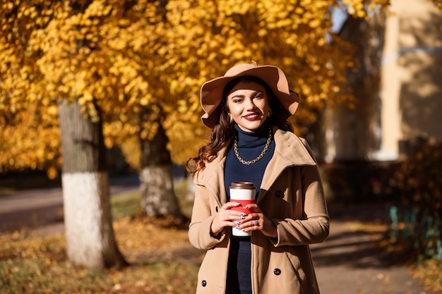 Modieuze vrouw in hoed met brede rand en beige jas houdt thermocup buiten in de herfst